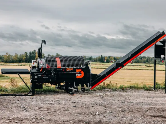En Palax D410 vedmaskin står utendørs på en grusplass med et jorde i bakgrunnen.