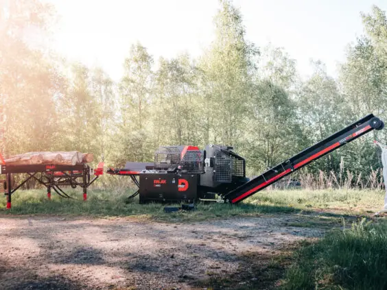 En Palax D460 vedmaskin med tømmerbord og transportbånd i et solfylt skogslandskap.