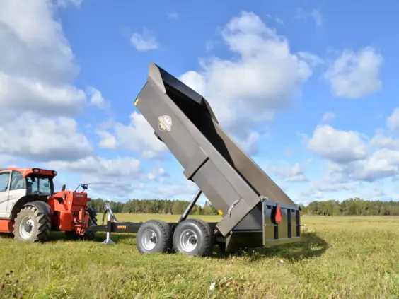Mørkegrå Palmse PT1000E dumperhenger tippet oppover, tilkoblet en rød traktor på et grønt jorde.