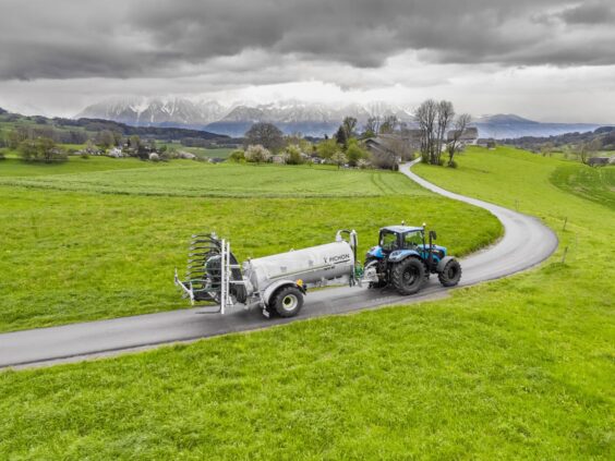 En blå traktor trekker en Pichon SV-serien vakumvogn langs en vei i et grønt landskap.