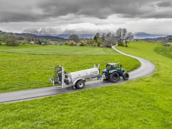 En blå traktor trekker en Pichon SV-serien vakumvogn langs en vei i et grønt landskap.