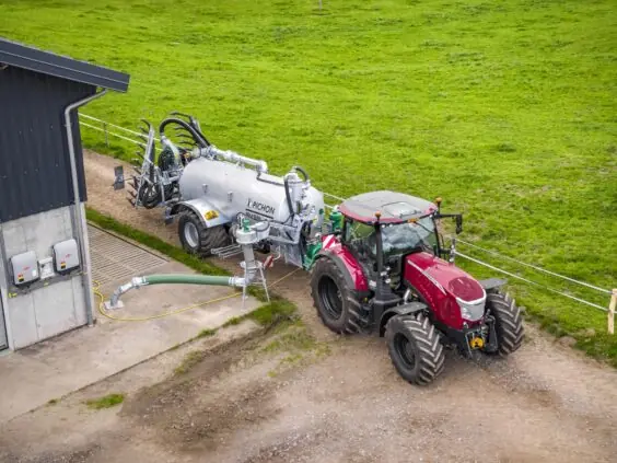 Rød traktor koblet til Pichon fyllesystemer for vakumvogner ved en landbruksbygning.