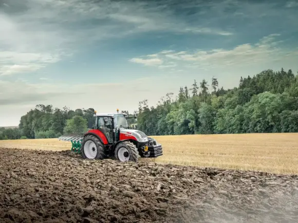 En rød og hvit Steyr Kompakt traktor som pløyer en åker under en overskyet himmel.