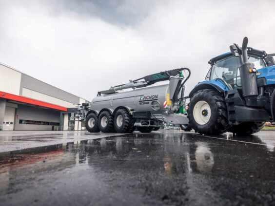 En blå traktor som trekker en grå Pichon SC24R gjødselvogn på våt asfalt.