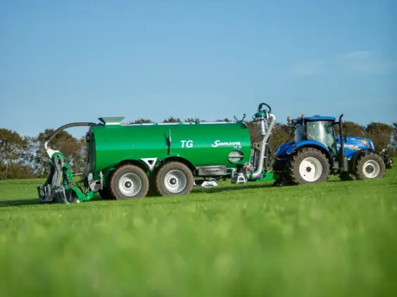En blå traktor som trekker en stor grønn gjødselvogn på en grønn eng.
