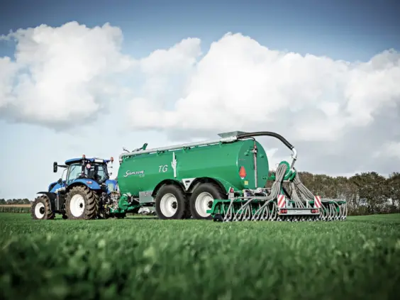 Blå traktor med grønn Samson gjødselspreder på en grønn mark under en skyete himmel.