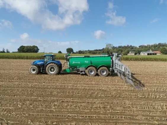 En blå traktor trekker en grønn Samson gjødselvogn på en stubbeåker.