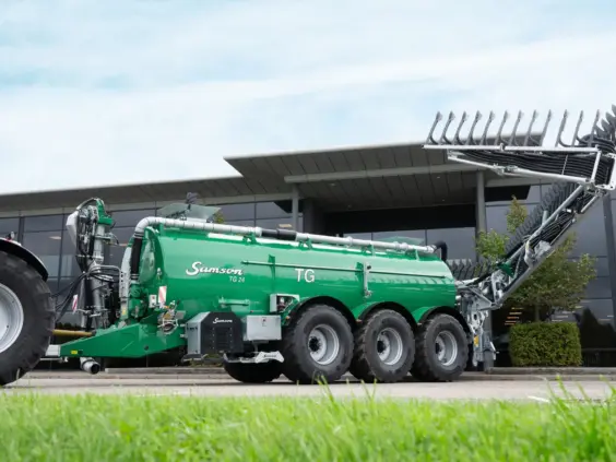 Samson TG 24 grønn gjødselvogn koblet til en traktor foran en moderne bygning.