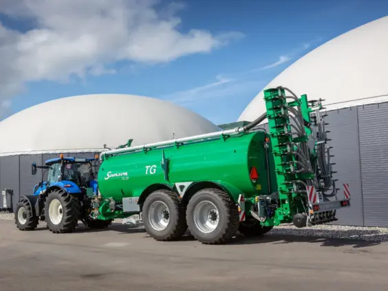 En blå traktor trekker en stor grønn Samson TG 20 gjødselvogn foran et biogassanlegg.