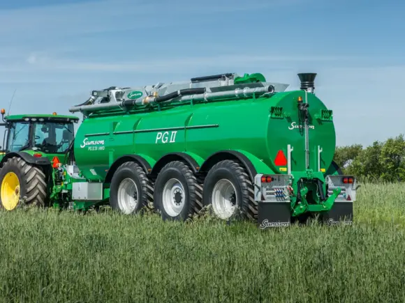 En grønn SAMSON PG II gjødselvogn trukket av en traktor i en grønn åker.