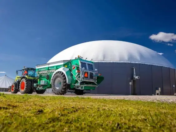 Traktor med samson Flex-II tørrgjødselvogner parkert foran et stort biogassanlegg under blå himmel.