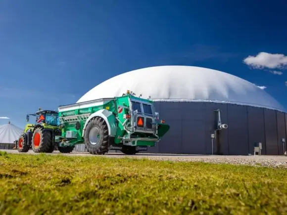 Traktor med samson Flex-II tørrgjødselvogner parkert foran et stort biogassanlegg under blå himmel.