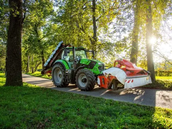 Grønn Deutz-Fahr Series 6 traktor med slåmaskin på en idyllisk vei med trær.