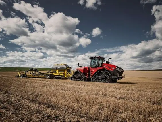 En rød Steiger 785 Quadtrac på belter trekker landbruksutstyr over en åker under blå himmel.