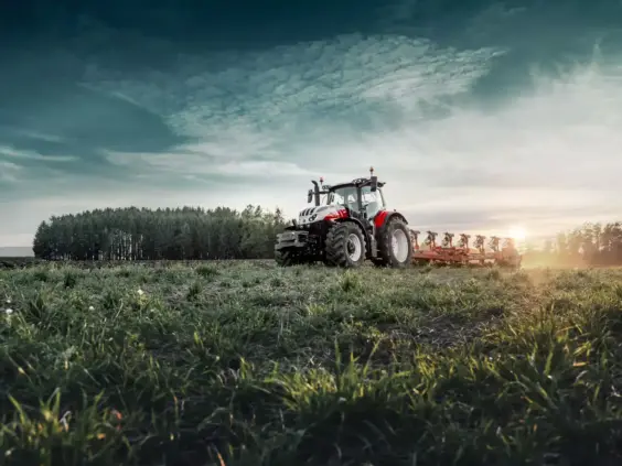 En rød og hvit Steyr Absolut CVT traktor pløyer en åker ved solnedgang.