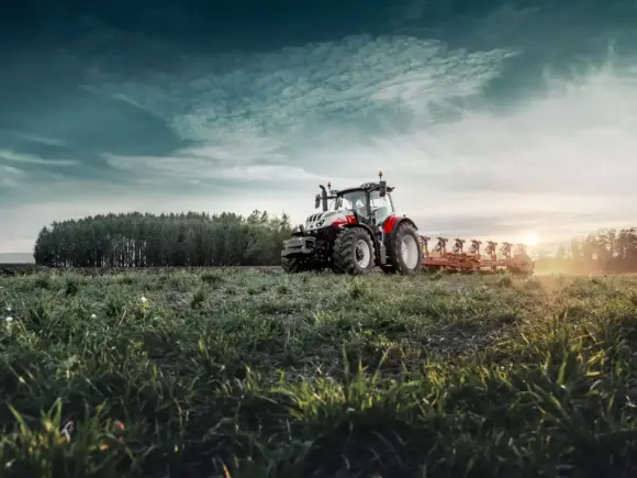 En rød og hvit Steyr Absolut CVT traktor pløyer en åker ved solnedgang.