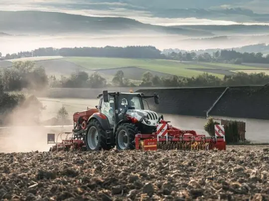 Rød og hvit Steyr Expert CVT traktor i arbeid på en åker i morgenlys.