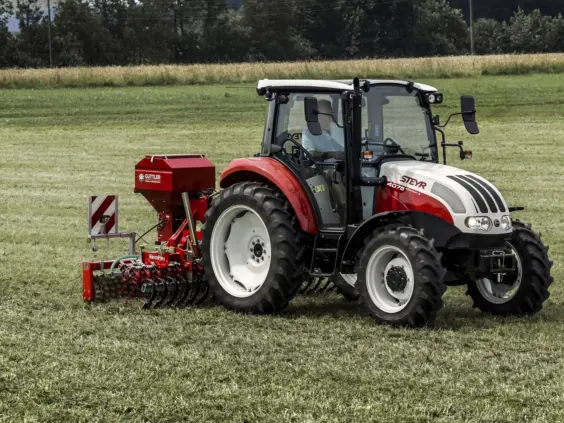 Steyr Kompakt S traktor med rødt landbruksutstyr i arbeid på en gresskledd mark.