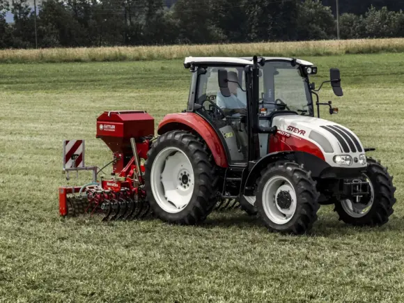 Steyr Kompakt S traktor med rødt landbruksutstyr i arbeid på en gresskledd mark.