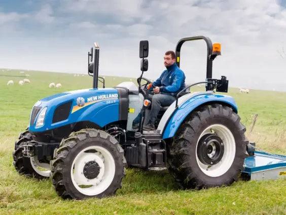 Blå New Holland T4S traktor i arbeid på et grønt jorde med sauer.