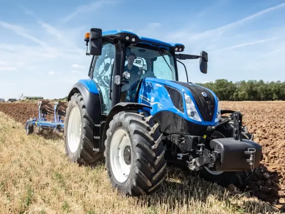 Blå New Holland traktor, T5 Dynamic Command og Auto Command, pløyer et jorde i solskinn.