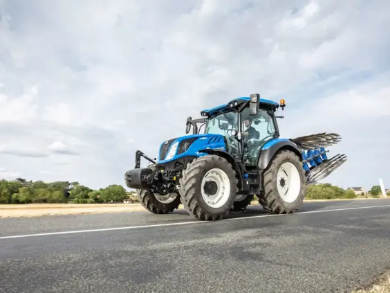Blå New Holland traktor, T5 Dynamic Command og Auto Command, kjører på vei med plog.