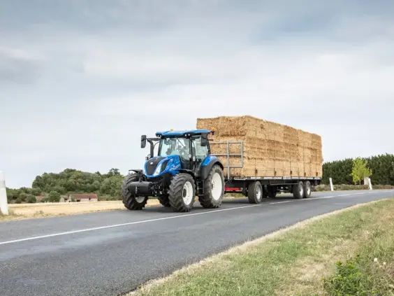Blå New Holland T5 Dynamic Command og Auto Command traktor med høyballer på en vei.