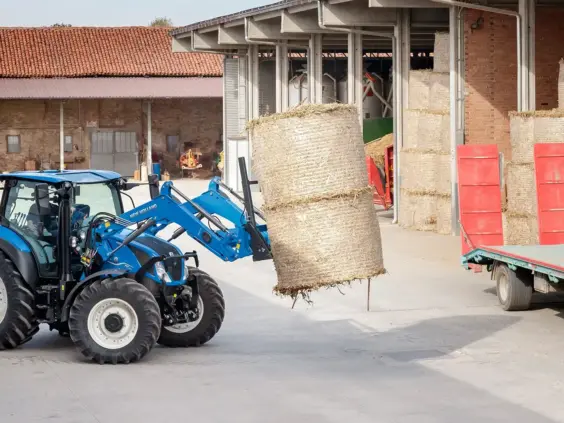 Blå New Holland T5 Dynamic Command og Auto Command traktor løfter høyball på gårdstunet.