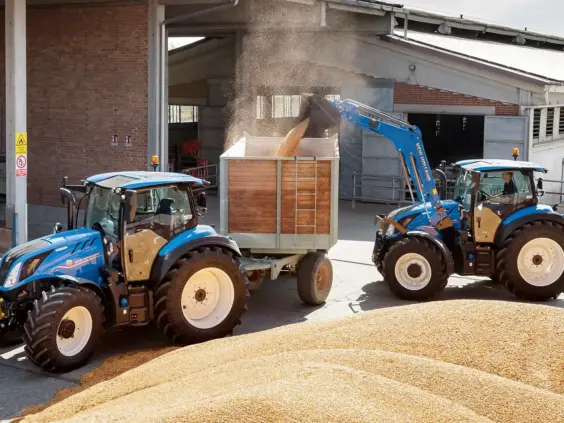 To blå New Holland-traktorer, T5 Dynamic Command og Auto Command, laster korn på en gård.