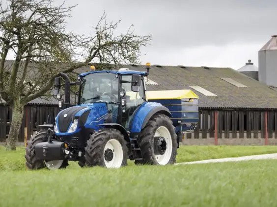 Blå New Holland T5 Electro Command traktor med gult utstyr foran en gårdsbygning.