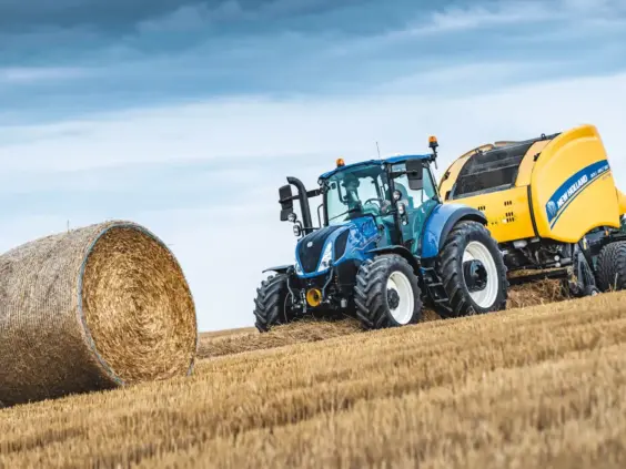 Blå New Holland T5 Electro Command traktor som presser høy på en gyllen åker.