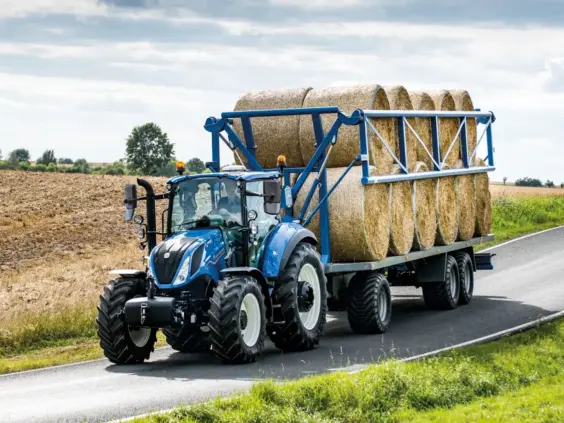 Blå New Holland T5 Electro Command traktor med tilhenger full av halmballer på en landevei.