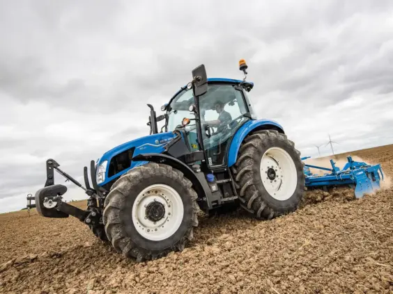 Blå New Holland T5 Utility traktor som pløyer en åker under en grå himmel.