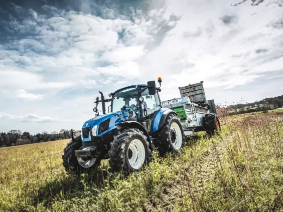 Blå New Holland T5 Utility traktor som sprer gjødsel på en åker under skyet himmel.