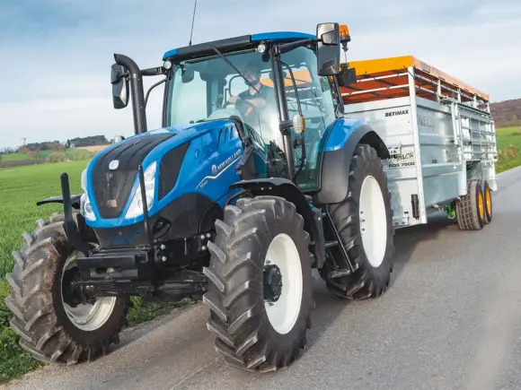 Blå New Holland T6 Electro / Dynamic / Auto Command traktor på vei med tilhenger.