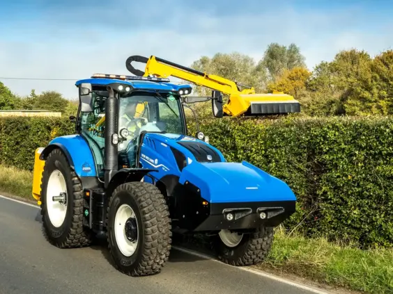 En blå New Holland T6.180 Methane Power traktor som klipper en hekk langs veien.