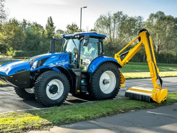 En blå New Holland T6.180 Methane Power traktor med gul kantklipper ved en vei.