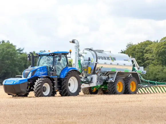 Blå New Holland T6.180 Methane Power traktor trekker en Joskin gjødselspreder på en åker.
