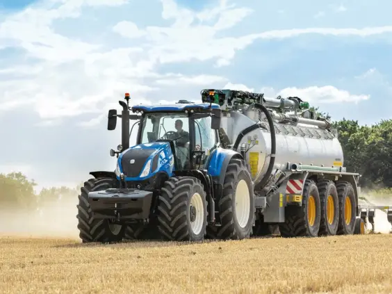 Blå New Holland T7 Heavy Duty traktor som trekker en stor tankvogn på et jorde.