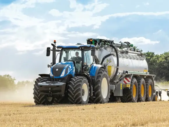 Blå New Holland T7 Heavy Duty traktor som trekker en stor tankvogn på et jorde.