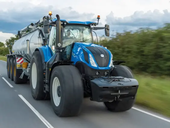 En blå New Holland T7 Heavy Duty traktor som trekker en stor tankvogn på veien.