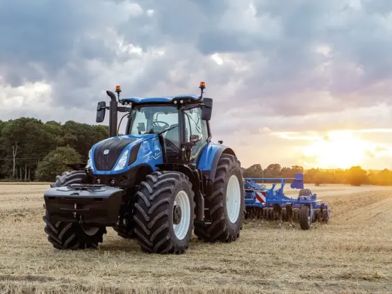 En blå New Holland T7 Heavy Duty traktor i arbeid på en åker ved solnedgang.