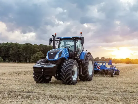 En blå New Holland T7 Heavy Duty traktor som arbeider i en åker ved solnedgang.