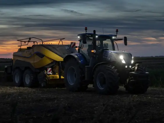 Blå traktor, T7 LWB med PLM Intelligence, i arbeid med ballepresse under en vakker solnedgang.