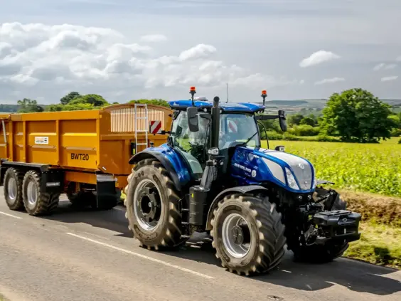 Blå New Holland T7 LWB med PLM Intelligence kjører med en gul tilhenger på vei.