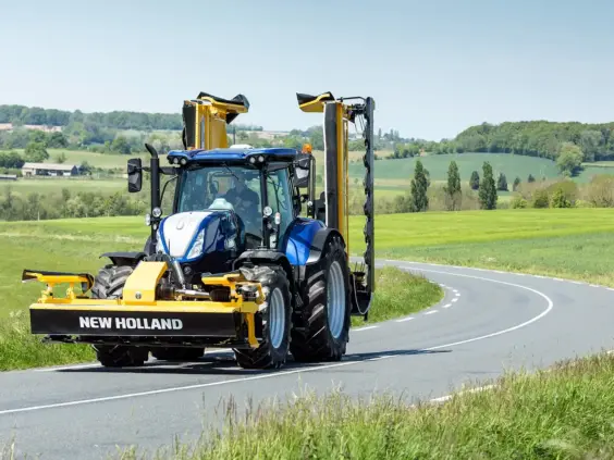 Blå New Holland T7 SWB traktor med slåmaskin på en svingete vei i landskapet.