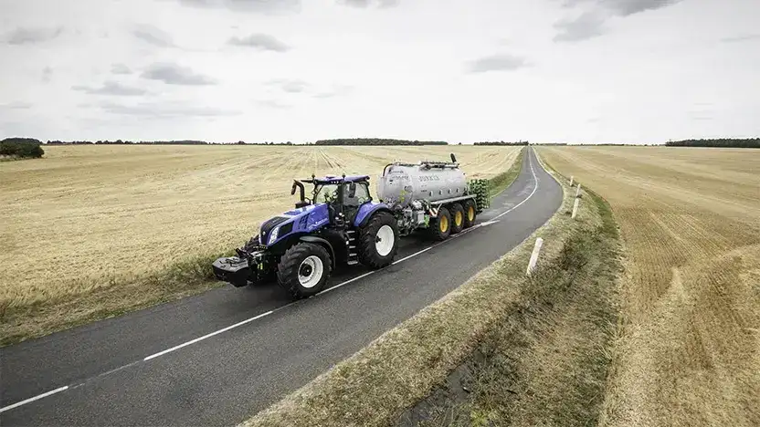 Blå New Holland T8 Genesis traktor med tankvogn på en landevei gjennom gule åkrer.