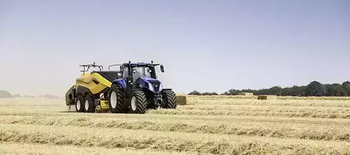 En blå New Holland T8 Genesis traktor trekker en gul ballepresse på et jorde.