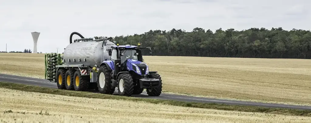 En blå New Holland T8 Genesis traktor som trekker en tankvogn langs et jorde.