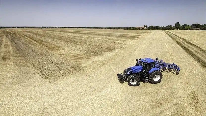 Blå New Holland T8 Genesis traktor som arbeider på et stort høstet jorde.